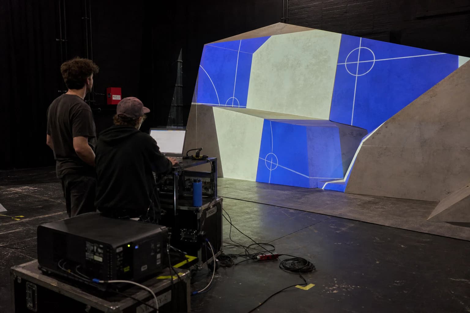 Zwei Techniker kalibrieren eine geometrische Bühnenfläche mit Projektor, Rechner und sichtbarer Verkabelung